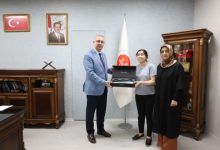 Görselde resmi bir makam odasında üç kişi yer alıyor. Bir kurum yöneticisi, ortadaki genç bir kadına kutu içerisinde bulunan bir cihazı veya hediyeyi takdim ederken poz veriyor. Yanlarında bulunan diğer kadın da törene eşlik ediyor. Arka planda Türk bayrağı, resmi kurum flaması ve devlet büyüklerine ait çerçeveli fotoğraflar bulunuyor. Fotoğraf, bir destek, ödül veya sosyal sorumluluk kapsamında gerçekleştirilen teslim törenini yansıtıyor.