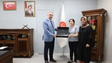 Görselde resmi bir makam odasında üç kişi yer alıyor. Bir kurum yöneticisi, ortadaki genç bir kadına kutu içerisinde bulunan bir cihazı veya hediyeyi takdim ederken poz veriyor. Yanlarında bulunan diğer kadın da törene eşlik ediyor. Arka planda Türk bayrağı, resmi kurum flaması ve devlet büyüklerine ait çerçeveli fotoğraflar bulunuyor. Fotoğraf, bir destek, ödül veya sosyal sorumluluk kapsamında gerçekleştirilen teslim törenini yansıtıyor.