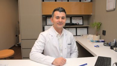 Fotoğrafta bir sağlık kuruluşundaki muayene odasında oturan bir uzman görülüyor. Beyaz doktor önlüğü ve açık renk gömlek giyen kişi, çalışma masasının arkasında kameraya dönük şekilde gülümsüyor. Arka planda çeşitli sertifika ve başarı belgelerinin yer aldığı raflar bulunuyor. Masanın üzerinde bilgisayar klavyesi, kalemler, evraklar ve küçük bir saksı bitkisi dikkat çekiyor. Ortam düzenli, aydınlık ve profesyonel bir klinik atmosferi yansıtıyor.