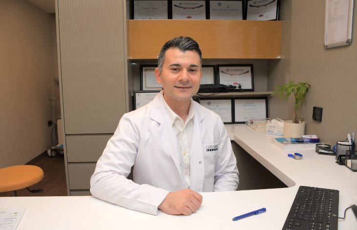 Fotoğrafta bir sağlık kuruluşundaki muayene odasında oturan bir uzman görülüyor. Beyaz doktor önlüğü ve açık renk gömlek giyen kişi, çalışma masasının arkasında kameraya dönük şekilde gülümsüyor. Arka planda çeşitli sertifika ve başarı belgelerinin yer aldığı raflar bulunuyor. Masanın üzerinde bilgisayar klavyesi, kalemler, evraklar ve küçük bir saksı bitkisi dikkat çekiyor. Ortam düzenli, aydınlık ve profesyonel bir klinik atmosferi yansıtıyor.