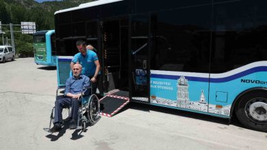 Görselde, tekerlekli sandalye kullanan bir kişi alçak tabanlı ve erişilebilir özelliklere sahip bir şehir otobüsünden inerken görülüyor. Bir görevli sandalyeyi yönlendirerek destek sağlıyor. Otobüsün girişinde engelli erişimine uygun rampa bulunurken, sahne toplu ulaşımda erişilebilirlik ve bağımsız hareket imkânını vurguluyor.