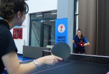 Görselde, kapalı bir spor salonunda masa tenisi maçı yapan iki sporcu yer alıyor. Ön planda sırtı dönük bir sporcu raketle topa vururken, karşı tarafta mavi forma giyen sporcu pozisyon almış şekilde oyuna odaklanıyor. Arka planda Türkiye Masa Tenisi Federasyonu'na ait bir afiş bulunuyor. Fotoğraf, antrenman veya müsabaka ortamını yansıtıyor.