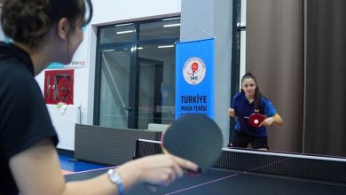Görselde, kapalı bir spor salonunda masa tenisi maçı yapan iki sporcu yer alıyor. Ön planda sırtı dönük bir sporcu raketle topa vururken, karşı tarafta mavi forma giyen sporcu pozisyon almış şekilde oyuna odaklanıyor. Arka planda Türkiye Masa Tenisi Federasyonu'na ait bir afiş bulunuyor. Fotoğraf, antrenman veya müsabaka ortamını yansıtıyor.