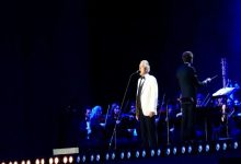 Görselde, bir sahnede mikrofon önünde performans sergileyen bir sanatçı ve arkasında yer alan senfoni orkestrası görülüyor. Mavi ışıklarla aydınlatılan sahnede, orkestrayı yöneten bir şef de yer alıyor. Fotoğraf, büyük bir konser organizasyonunda gerçekleşen canlı müzik performansından bir anı yansıtıyor.