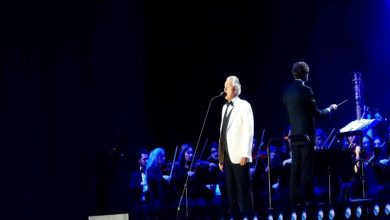 Görselde, bir sahnede mikrofon önünde performans sergileyen bir sanatçı ve arkasında yer alan senfoni orkestrası görülüyor. Mavi ışıklarla aydınlatılan sahnede, orkestrayı yöneten bir şef de yer alıyor. Fotoğraf, büyük bir konser organizasyonunda gerçekleşen canlı müzik performansından bir anı yansıtıyor.