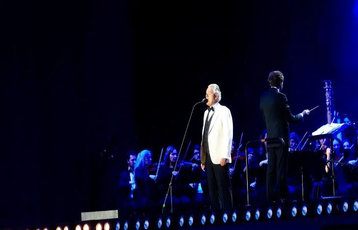 Görselde, bir sahnede mikrofon önünde performans sergileyen bir sanatçı ve arkasında yer alan senfoni orkestrası görülüyor. Mavi ışıklarla aydınlatılan sahnede, orkestrayı yöneten bir şef de yer alıyor. Fotoğraf, büyük bir konser organizasyonunda gerçekleşen canlı müzik performansından bir anı yansıtıyor.