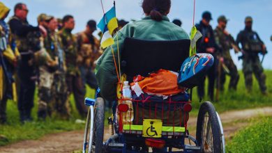 Tekerlekli sandalyedeki bir kişi, arkasında engellilik simgesi ve küçük bayraklar bulunan aracıyla, karşısında kamuflajlı askerlerin durduğu açık bir alanda görülüyor. Gökyüzü bulutlu, ortam kırsal bir yol kenarı.