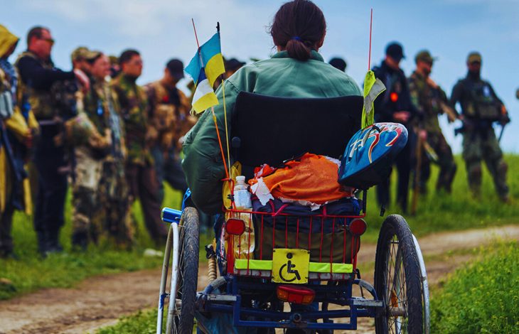 Tekerlekli sandalyedeki bir kişi, arkasında engellilik simgesi ve küçük bayraklar bulunan aracıyla, karşısında kamuflajlı askerlerin durduğu açık bir alanda görülüyor. Gökyüzü bulutlu, ortam kırsal bir yol kenarı.