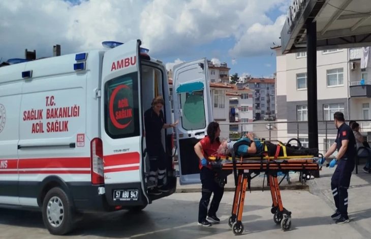 Bir ambulansın yanında sağlık ekiplerinin sedye üzerindeki yaralı bir kişiye müdahale ettiği görülüyor. Acil sağlık personeli, yaralıyı ambulansa taşımaya hazırlanırken olayın bir sağlık kuruluşu veya hastane çevresinde gerçekleştiği anlaşılıyor. Görsel, trafik kazası sonrası yürütülen acil müdahale sürecini yansıtıyor.