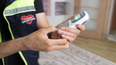 Görselde bir görevlinin elinde taşınabilir bir elektronik cihaz tuttuğu görülüyor. Üzerindeki yelekte yer alan kalp logosu ve cihazın yapısı, sağlık veya sosyal destek hizmetleri kapsamında kullanılan bir ekipmanı işaret ediyor. Fotoğraf, ev ortamında gerçekleştirilen bir hizmet veya ziyaret sırasında çekilmiş izlenimi veriyor.
