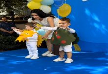 Mavi zeminli bir sahnede, kostüm giymiş iki küçük çocuk ve yanlarında onları yönlendiren bir kadın görülüyor. Çocuklardan biri güneş, diğeri ise ağaç temalı kostüm taşırken, etkinlik alanı sarı-beyaz-mavi balonlarla süslenmiş. Çocuklar sahnede neşeli bir şekilde izleyicilere doğru poz veriyor.
