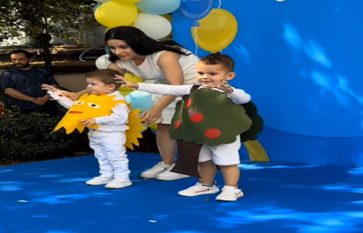 Mavi zeminli bir sahnede, kostüm giymiş iki küçük çocuk ve yanlarında onları yönlendiren bir kadın görülüyor. Çocuklardan biri güneş, diğeri ise ağaç temalı kostüm taşırken, etkinlik alanı sarı-beyaz-mavi balonlarla süslenmiş. Çocuklar sahnede neşeli bir şekilde izleyicilere doğru poz veriyor.