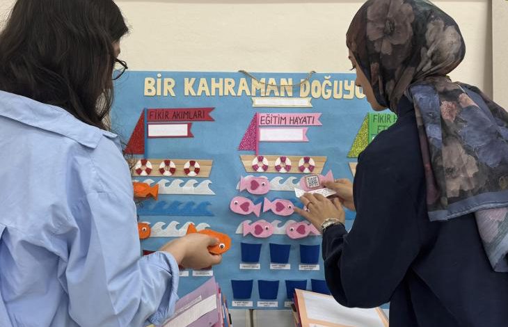 İki kişi renkli bir eğitim panosu önünde duruyor, kâğıt balıklarla etkileşimli bir çalışma yapıyor.