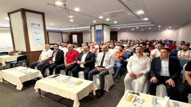 Görselde konferans veya seminer salonunda düzenlenen bir etkinlik görülüyor. Ön sırada takım elbiseli ve resmi kıyafetli katılımcılar otururken, arka sıralarda kalabalık bir dinleyici grubu yer alıyor. Salonda projeksiyon cihazı ve toplantı düzeni dikkat çekiyor; ortam eğitim ve bilgilendirme toplantısı atmosferi taşıyor.