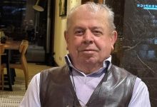 Görselde iç mekânda çekilmiş bir portre yer alıyor. Bir kişi kameraya dönük şekilde oturuyor; açık renk gömlek ve koyu renk deri yelek giyiyor. Arka planda masa, sandalyeler ve sıcak ışıklı bir ortam görülüyor. Fotoğraf sakin ve samimi bir anı yansıtıyor.