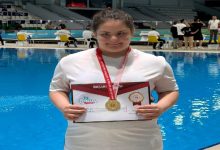 Bir yüzme havuzunun önünde çekilen fotoğrafta, boynunda altın madalya taşıyan genç bir sporcu başarı belgesini tutarak poz veriyor. Beyaz tişört giyen sporcu kameraya gülümserken, arka planda havuz ve müsabaka alanında bulunan görevliler görülüyor. Fotoğraf, yüzme yarışmasında elde edilen birincilik başarısını yansıtıyor.