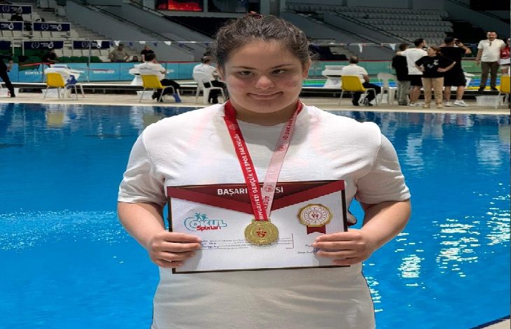 Bir yüzme havuzunun önünde çekilen fotoğrafta, boynunda altın madalya taşıyan genç bir sporcu başarı belgesini tutarak poz veriyor. Beyaz tişört giyen sporcu kameraya gülümserken, arka planda havuz ve müsabaka alanında bulunan görevliler görülüyor. Fotoğraf, yüzme yarışmasında elde edilen birincilik başarısını yansıtıyor.