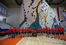Kapalı bir spor salonundaki yüksek yapay tırmanış duvarının önünde, kırmızı formalar ve emniyet kemerleri giymiş Para Tırmanış Milli Takımı sporcuları toplu halde poz veriyor.