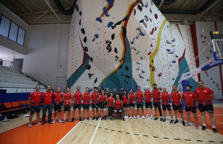 Kapalı bir spor salonundaki yüksek yapay tırmanış duvarının önünde, kırmızı formalar ve emniyet kemerleri giymiş Para Tırmanış Milli Takımı sporcuları toplu halde poz veriyor.