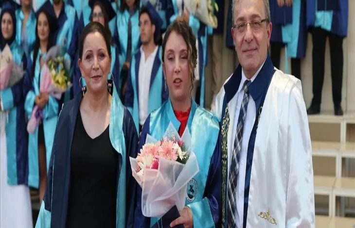 Görselde, mezuniyet töreninde çekilmiş bir an yer alıyor. Ortada mezun cübbesi giymiş genç bir kadın elinde çiçek buketiyle dururken, yanında annesi ve akademik cübbe giymiş bir öğretim üyesi poz veriyor. Arka planda ise törene katılan diğer mezunlar görülüyor.