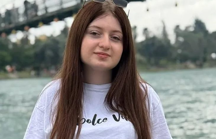 Fotoğrafta uzun kahverengi saçlı genç bir kız, göl kenarında objektife bakarak poz veriyor. Arka planda su, ağaçlar ve bir köprü yer alıyor.