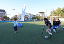 Suni çim sahada koltuk değnekleriyle futbol oynayan engelli sporcular antrenman yapıyor. Arka planda diğer sporcular ve etkinliği takip eden katılımcılar görülüyor.