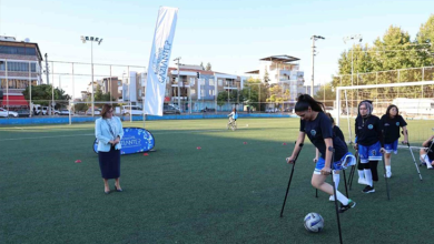 Suni çim sahada koltuk değnekleriyle futbol oynayan engelli sporcular antrenman yapıyor. Arka planda diğer sporcular ve etkinliği takip eden katılımcılar görülüyor.