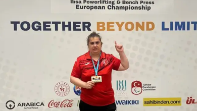 Görme engelli milli halterci, boynunda madalyasıyla Avrupa Şampiyonası'nın organizasyon panosu önünde poz veriyor. Üzerinde Türkiye formasını taşıyan sporcu, elde ettiği başarının ardından objektife işaret parmağını kaldırarak poz veriyor.