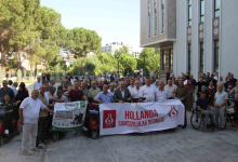 Bir kamu binasının önünde toplanan kalabalık, elektrikli engelli araçlarıyla birlikte hatıra fotoğrafı çektiriyor. Ön sırada yardım kampanyasına ait pankartlar taşınırken, etkinliğe katılan engelli bireyler ve destekçiler bir arada görülüyor.