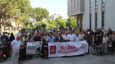 Bir kamu binasının önünde toplanan kalabalık, elektrikli engelli araçlarıyla birlikte hatıra fotoğrafı çektiriyor. Ön sırada yardım kampanyasına ait pankartlar taşınırken, etkinliğe katılan engelli bireyler ve destekçiler bir arada görülüyor.
