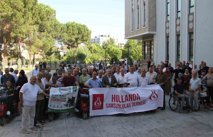 Bir kamu binasının önünde toplanan kalabalık, elektrikli engelli araçlarıyla birlikte hatıra fotoğrafı çektiriyor. Ön sırada yardım kampanyasına ait pankartlar taşınırken, etkinliğe katılan engelli bireyler ve destekçiler bir arada görülüyor.
