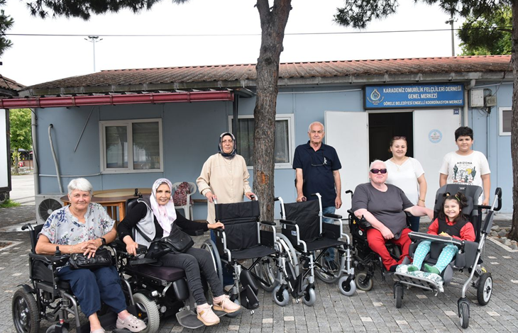 Karadeniz Omurilik Felçlileri Derneği önünde çekilen fotoğrafta, tekerlekli sandalye ve akülü sandalye kullanan bireyler ile yakınları, teslim edilen tekerlekli sandalyelerle birlikte toplu halde objektife poz veriyor. Arka planda dernek binasının girişi ve tabelası yer alıyor.