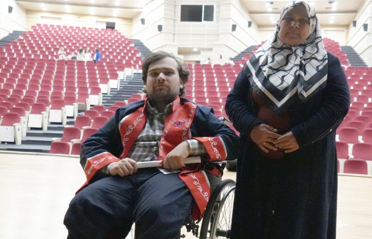 Abdulsamet Sert, mezuniyet cübbesi giyinmiş olarak tekerlekli sandalyede bulunuyor ve annesi yanında ayakta durmakta.