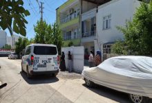 Görselde, bir apartmanın önünde park halindeki polis aracı ile bina girişinde bekleyen birkaç kişi görülüyor. Sokakta üzeri branda ile örtülü bir otomobil yer alırken, olay yerine polis ekiplerinin geldiği ve çevredeki vatandaşların gelişmeleri izlediği an yansıyor.