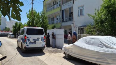 Görselde, bir apartmanın önünde park halindeki polis aracı ile bina girişinde bekleyen birkaç kişi görülüyor. Sokakta üzeri branda ile örtülü bir otomobil yer alırken, olay yerine polis ekiplerinin geldiği ve çevredeki vatandaşların gelişmeleri izlediği an yansıyor.