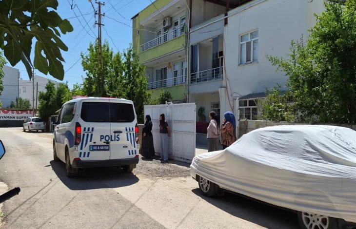 Görselde, bir apartmanın önünde park halindeki polis aracı ile bina girişinde bekleyen birkaç kişi görülüyor. Sokakta üzeri branda ile örtülü bir otomobil yer alırken, olay yerine polis ekiplerinin geldiği ve çevredeki vatandaşların gelişmeleri izlediği an yansıyor.