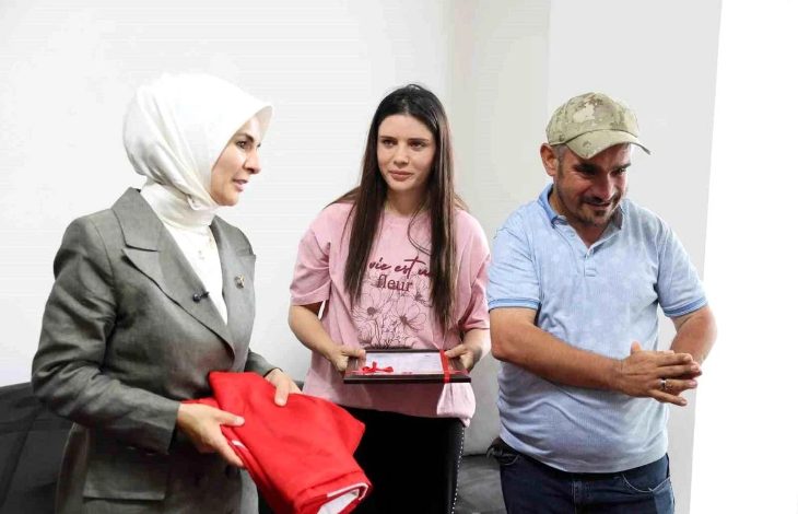Görselde, Aile ve Sosyal Hizmetler Bakanı Mahinur Özdemir Göktaş, pembe tişörtlü genç bir kadın ve şapkalı bir erkek aynı ortamda ayakta duruyor. Genç kadın elinde çerçeveli bir hediye tutarken, Bakan katlanmış kırmızı bir kumaş taşıyor. Üçlü gülümseyerek sohbet ediyor ve görüntü, samimi bir ziyaret sırasında hediye takdimi yapılan anı yansıtıyor.