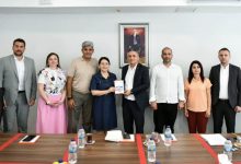 Toplantı salonunda, uzun bir masanın arkasında yedi kişi yan yana duruyor. Masanın üzerinde su şişeleri, not defterleri ve çay bardakları bulunuyor. Ortada yer alan iki kişi, Ankara Kent Konseyi Engelli Meclisi tarafından hazırlanan raporu temsil eden bir dosyayı birlikte tutarak poz veriyor. Katılımcıların tamamı resmi veya yarı resmi kıyafetler içinde, toplantı atmosferine uygun bir duruş sergiliyor. Arka duvarda Mustafa Kemal Atatürk’ün çerçeveli portresi yer alıyor. Genel ortam; kurumsal, ciddi ve iş birliğine dayalı bir görüşme izlenimi veriyor.