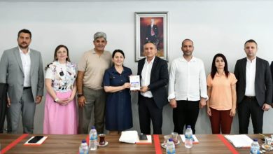 Toplantı salonunda, uzun bir masanın arkasında yedi kişi yan yana duruyor. Masanın üzerinde su şişeleri, not defterleri ve çay bardakları bulunuyor. Ortada yer alan iki kişi, Ankara Kent Konseyi Engelli Meclisi tarafından hazırlanan raporu temsil eden bir dosyayı birlikte tutarak poz veriyor. Katılımcıların tamamı resmi veya yarı resmi kıyafetler içinde, toplantı atmosferine uygun bir duruş sergiliyor. Arka duvarda Mustafa Kemal Atatürk’ün çerçeveli portresi yer alıyor. Genel ortam; kurumsal, ciddi ve iş birliğine dayalı bir görüşme izlenimi veriyor.