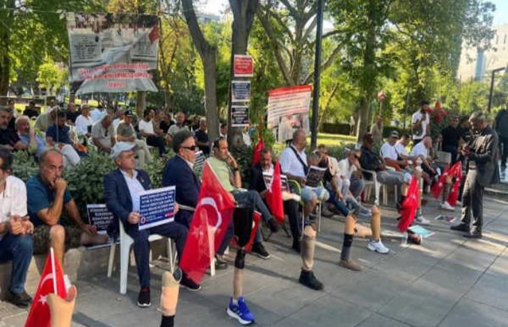 Parkta toplanan gaziler, ellerinde Türk bayrakları ve çeşitli pankartlarla oturarak bir protesto etkinliğine katılıyor; bazı katılımcıların protez bacak kullandığı da görülüyor.