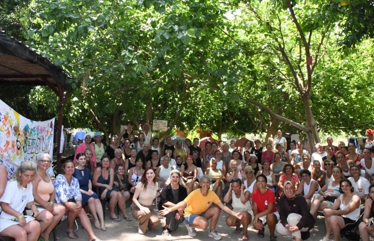 Görselde, ağaçların gölgesindeki açık alanda çok sayıda kişinin bir araya geldiği toplu bir etkinlik görülüyor. Katılımcılar kameraya gülümseyerek poz verirken, ön tarafta çömelmiş ve oturan kişiler, arka tarafta ise ayakta duran kalabalık yer alıyor.