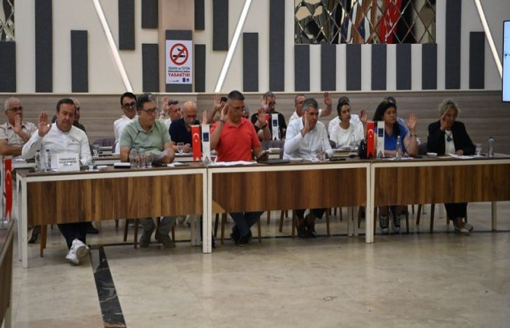 Görselde, uzun bir toplantı masası etrafında oturan belediye meclis üyelerinin el kaldırarak oylama yaptığı an yer alıyor. Masaların üzerinde Türk bayrakları, mikrofonlar ve evraklar bulunurken, toplantı salonundaki resmi ve karar alma süreci dikkat çekiyor.