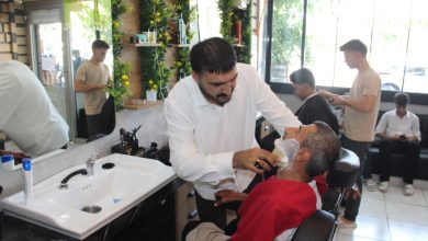 Görselde bir berber, tekerlekli sandalyede oturan yaşlı bir müşteriye sakal tıraşı yapıyor. Aydınlık bir kuaför salonunda çekilen fotoğrafta, arka planda diğer müşteriler ve çalışanlar günlük işlerini sürdürürken görülüyor.