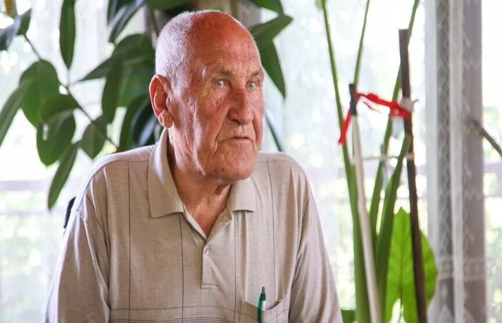 Görselde, açık renk polo tişört giyen yaşlı bir adam iç mekânda otururken kameraya doğru ciddi bir ifadeyle bakıyor. Arka planda büyük yeşil yapraklı saksı bitkileri ve gün ışığı alan bir pencere bulunuyor. Portre niteliğindeki görüntü, röportaj veya söyleşi ortamını çağrıştırıyor.