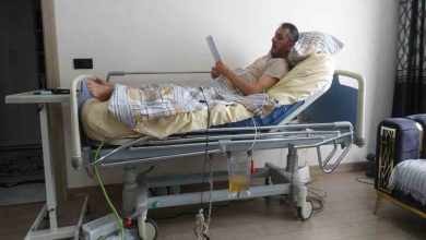 Mehmet Ali Arslan, elindeki belgeyi incelerken tıbbi ekipmanlarla donatılmış bir hasta yatağında dinlenmektedir.