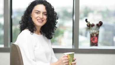Görselde, bir ofis ortamında oturan ve elinde bir kupa tutan kadın kameraya gülümseyerek poz veriyor. Arka planda geniş pencerelerden şehir manzarası görülürken, pencere önünde dekoratif çiçekler bulunuyor. Sade ve aydınlık ofis ortamı profesyonel bir röportaj atmosferi yansıtıyor.