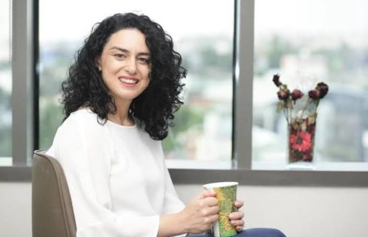 Görselde, bir ofis ortamında oturan ve elinde bir kupa tutan kadın kameraya gülümseyerek poz veriyor. Arka planda geniş pencerelerden şehir manzarası görülürken, pencere önünde dekoratif çiçekler bulunuyor. Sade ve aydınlık ofis ortamı profesyonel bir röportaj atmosferi yansıtıyor.