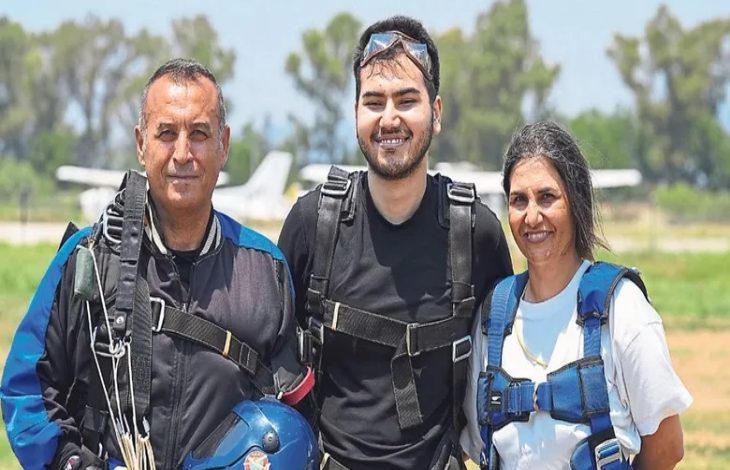 Görselde, açık alanda paraşüt atlayışı öncesinde çekilmiş bir hatıra karesi yer alıyor. Ortada paraşüt ekipmanlarını kuşanmış genç bir kişi, iki eğitmenle birlikte objektife gülümseyerek poz veriyor. Üçünün de üzerinde tandem atlayışa uygun emniyet kemerleri bulunurken, arka planda pist ve küçük uçaklar görülüyor.