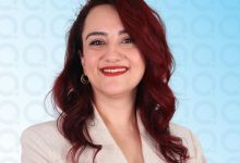 Koyu kızıl dalgalı saçlı, açık renk blazer giyen, altın halka küpeli; mavi tonlu modern bir arka planda kameraya sakin ve profesyonel bir ifadeyle bakan bir portre.