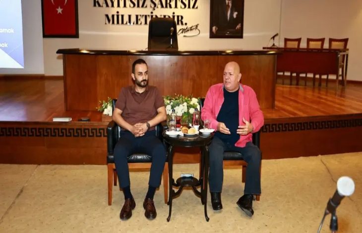 Görselde, bir belediye meclis salonunda düzenlenen eğitim programında iki konuşmacı dinleyicilere hitap ediyor. Ön tarafta yan yana oturan konuşmacıların arasında küçük bir masa bulunurken, arka planda "Kayıtsız Şartsız Milletindir" yazısı, Türk bayrağı ve Atatürk portresi yer alıyor. Eğitim veya söyleşi ortamı dikkat çekiyor.